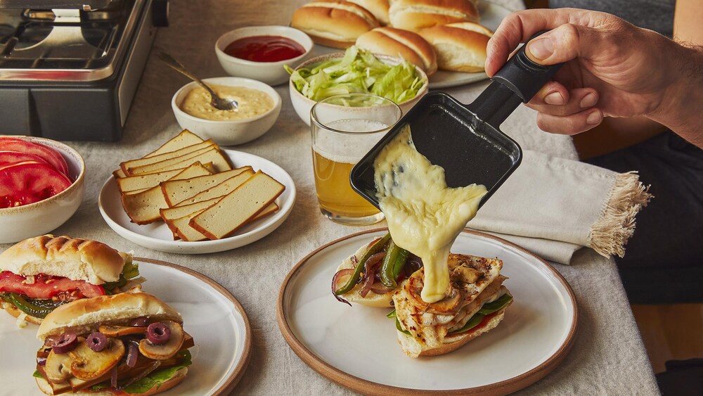 Dans une assiette, il y a deux petits sous-marins garnis différemment. À côté, une personne verse du fromage fondu de sa coupelle à son sous-marin. Autour, il y a une assiette de tofu fumé, deux bols de sauce, un bol de laitue et une assiette pleine de petits pains à salade.