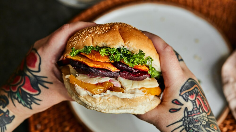 Il y a deux mains qui tiennent le sandwich et en arrière plan, il est possible de voir une assiette sur un napperon. 