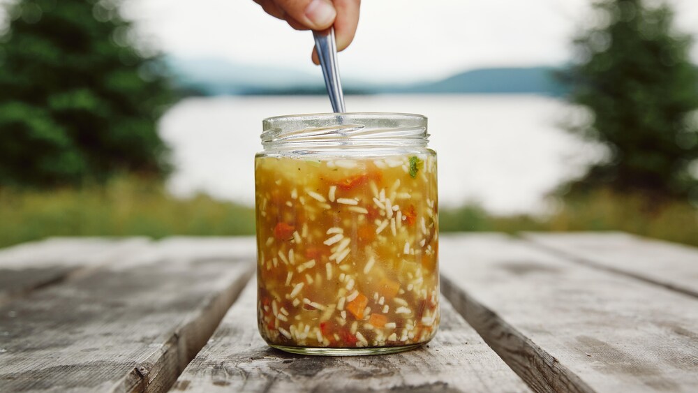 Un pot en verre, contenant de la soupe au jerky et au riz, est déposé sur une table en bois. Un main est au-dessus du pot en verre et elle tient la cuillère qui est dans le pot.
