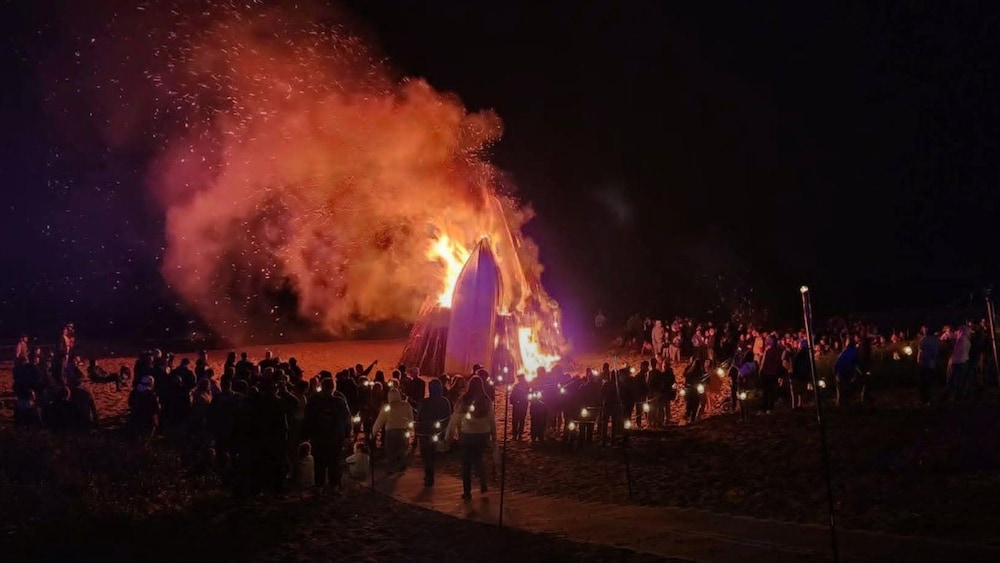 Foule rassemblé devant un feu dans lequel brûle des épaves de bateaux.