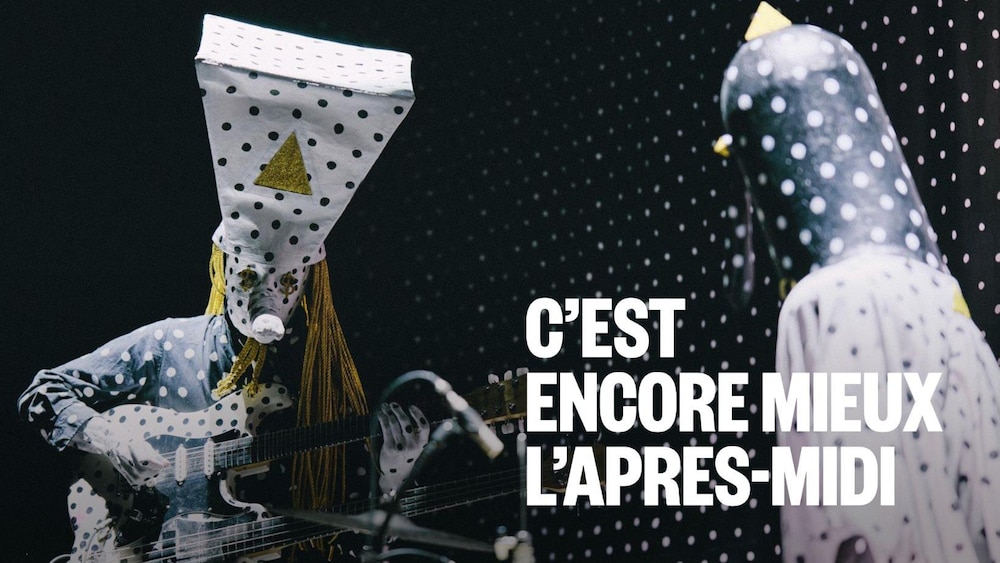 Deux musiciens masqués et vêtus de tenues à pois noirs et blancs performent sur scène. L'un joue d'une guitare double manche sous une coiffe trapézoïdale imposante, tandis que l'autre, de profil, s'installe derrière une batterie devant un fond pointillé.