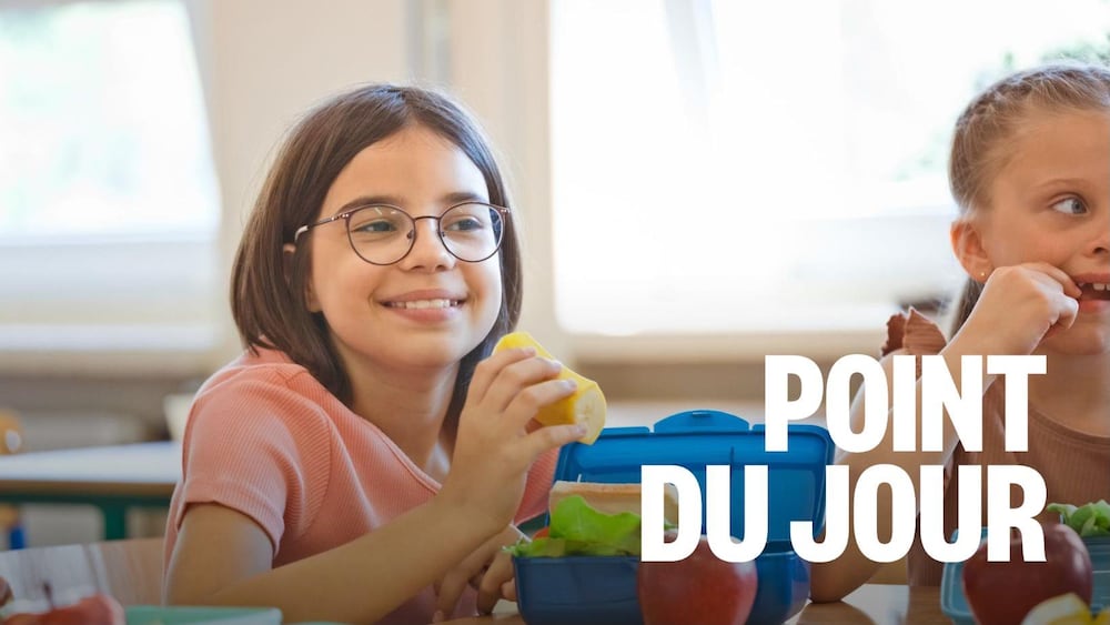 Une petite fille portant des lunettes tient un morceau de banane au-dessus de sa boîte à lunch ouverte sur la table.
