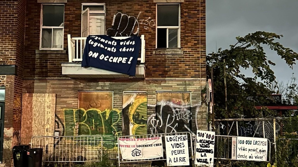 Un triplex abandonné. Sur le balcon, une banderole avec l'inscription « Logements vides, logements chers, on occupe! ». Une clôture devant l'immeuble pour empêcher d'accéder au terrain.