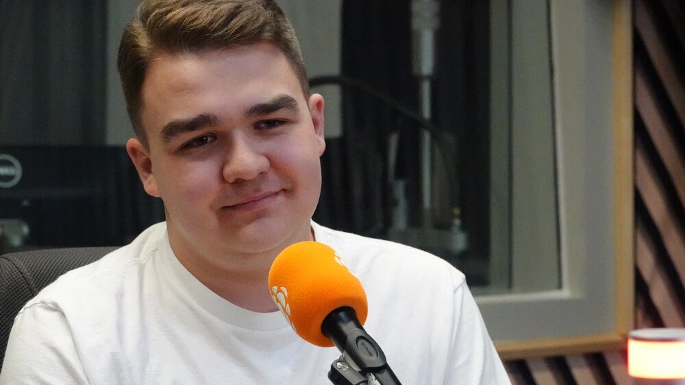 Un jeune homme souriant portant des lunettes et un chandail blanc, s'exprimant devant un microphone.