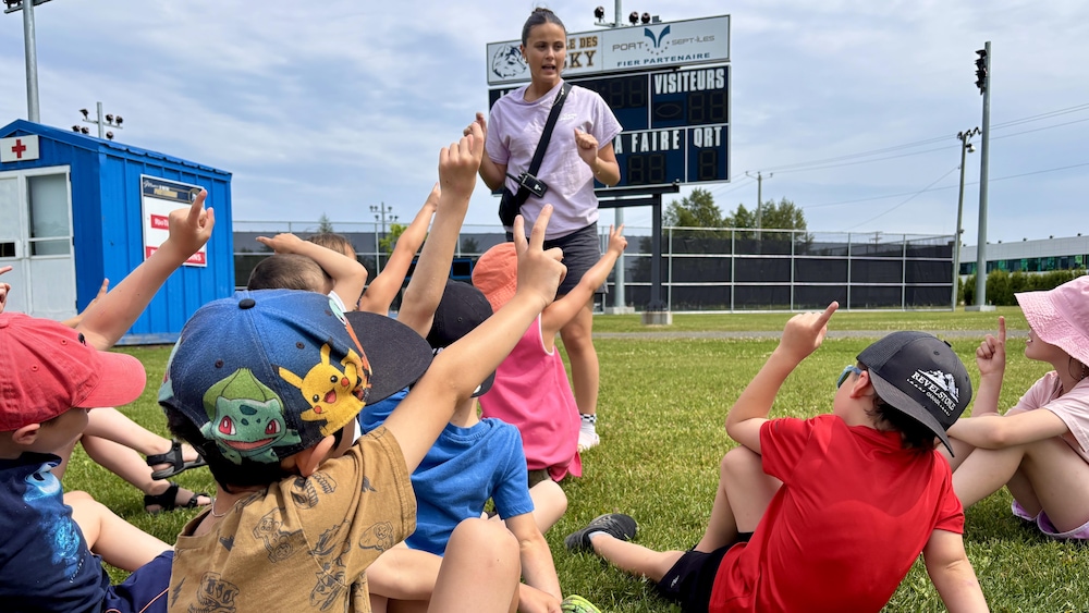 Julianne Lynch s'adresse à un groupe de jeunes enfants assis dans le gazon, au camp de jour de Sept-Îles. Ils ont tous la main levée, avec un ou deux doigts dressés, pour indiquer leur équipe.