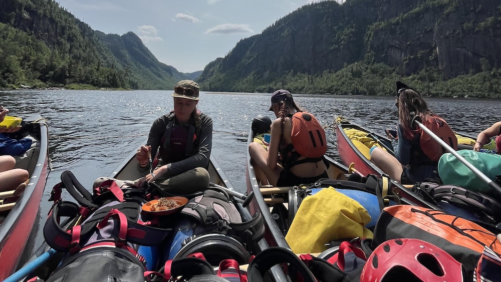 De jeunes canoteurs dînent dans leurs canots. Ils se trouvent sur la rivière Moisie, bordée par d'immenses falaises.