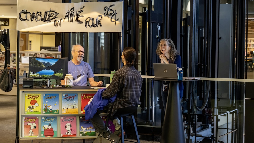 Hervé Simoneau, assis derrière un kiosque, rit de bon coeur avec un jeune homme qui le consulte, sous le regard hilare de Caroline Malo un peu en périphérie.