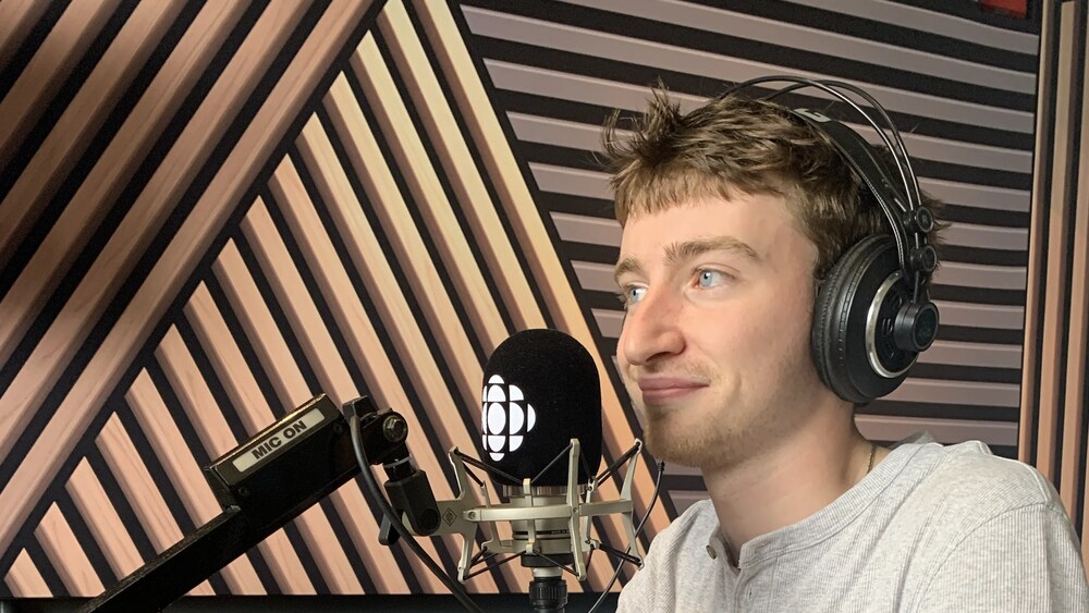 Un jeune homme portant un casque d'écoute est assis devant un micro et une tasse aux couleurs de Radio-Canada dans un studio de radio.