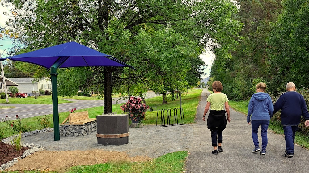 Trois aînés marchent sur un sentier en nature près d'une rue, un banc et un parasol.