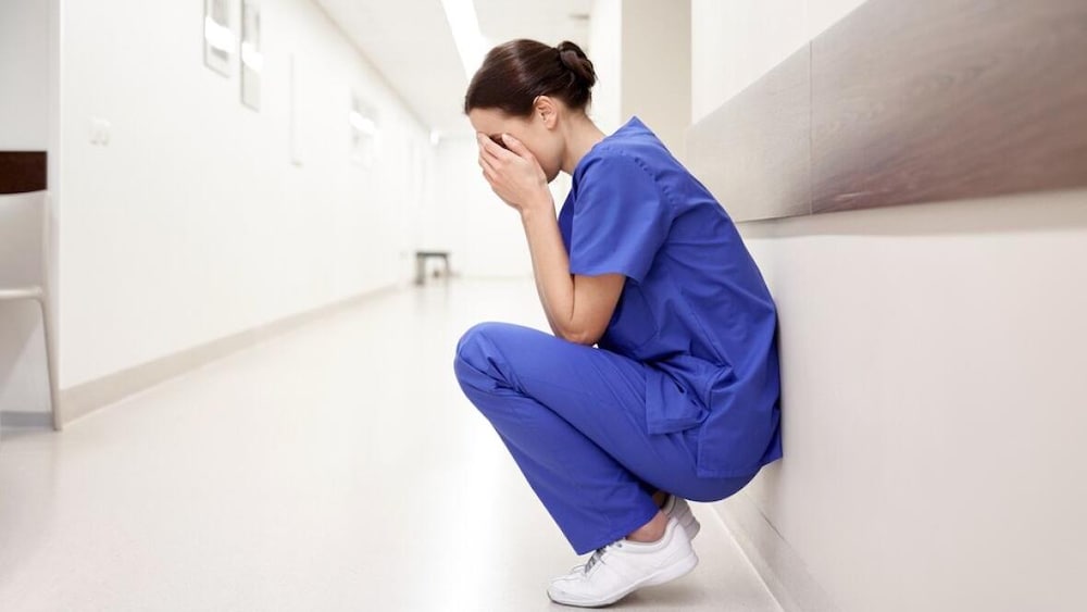 Une infirmière repliée sur elle-même dans un corridor d'hôpital, le visage dans les mains. 