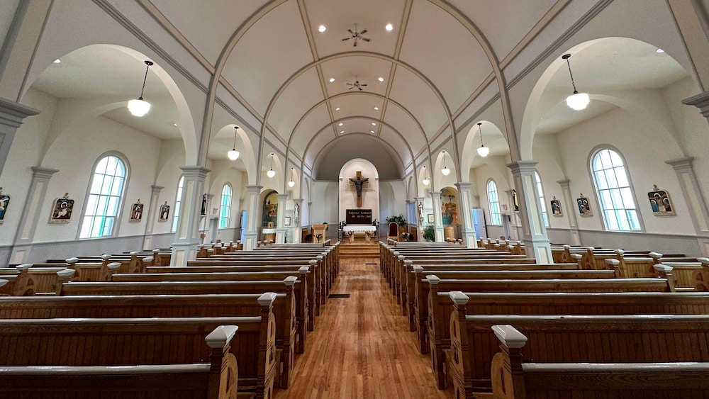 Photo de l'intérieur de l'église prise de l'allée centrale, entre deux rangées de bancs, et avec vue sur l'autel au loin.