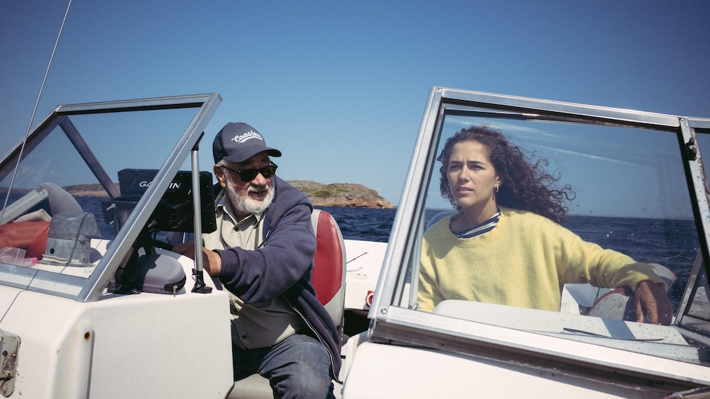 Plan sur Gilles Monger et Julia Pagé, à bord d'un bateau à moteur. Ils se trouvent en pleine mer, au cœur de l'archipel de Tête-à-la-Baleine. Le temps est clément, une brise agite leurs cheveux.
