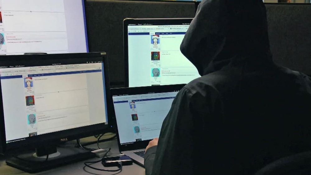Un homme en capuche devant une série d'ordinateurs sur lesquels sont ouvertes des pages de forums de discussion.