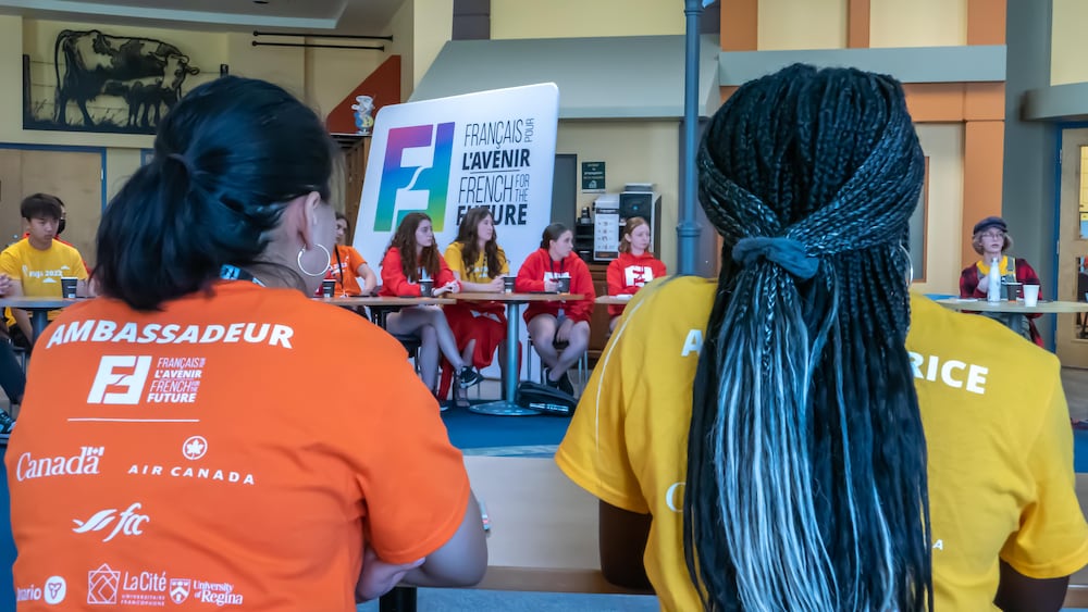 Vue arrière de deux jeunes participants. L'un porte un chandail orange avec l'inscription « AMBASSADEUR » et un logo du Canada, l'autre a de longues tresses bleues et porte un chandail jaune.AMBASSADEUR » et un logo du Canada, l'autre a de longues tresses bleues et porte un chandail jaune.