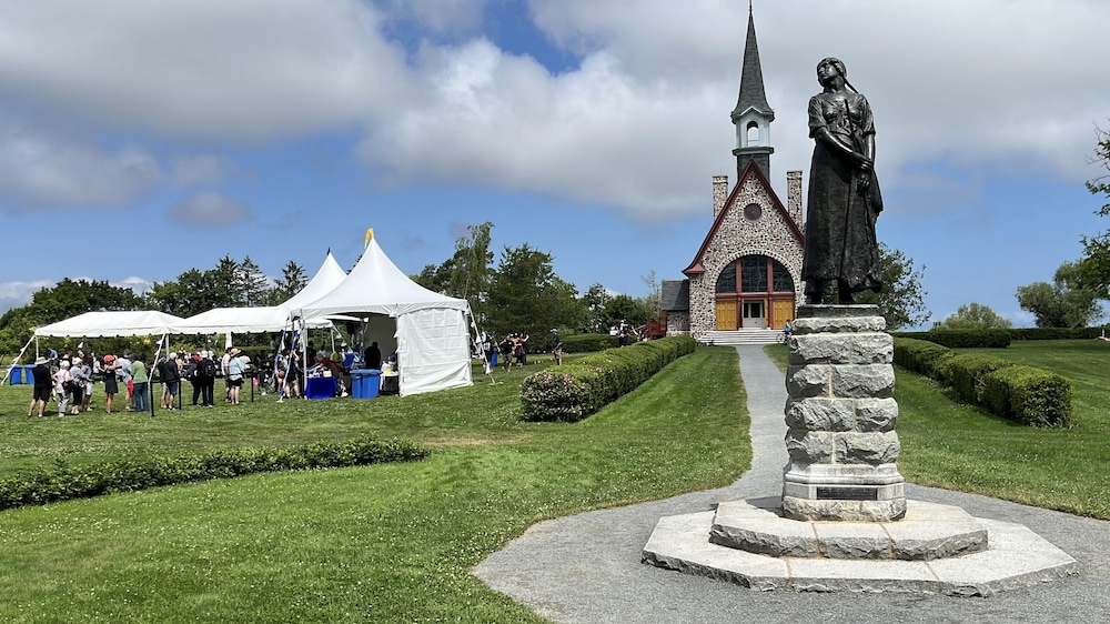 La statue d'Évangéline, une église et, à gauche de la photo, un chapiteau blanc avec des dizaines de personnes dessous.