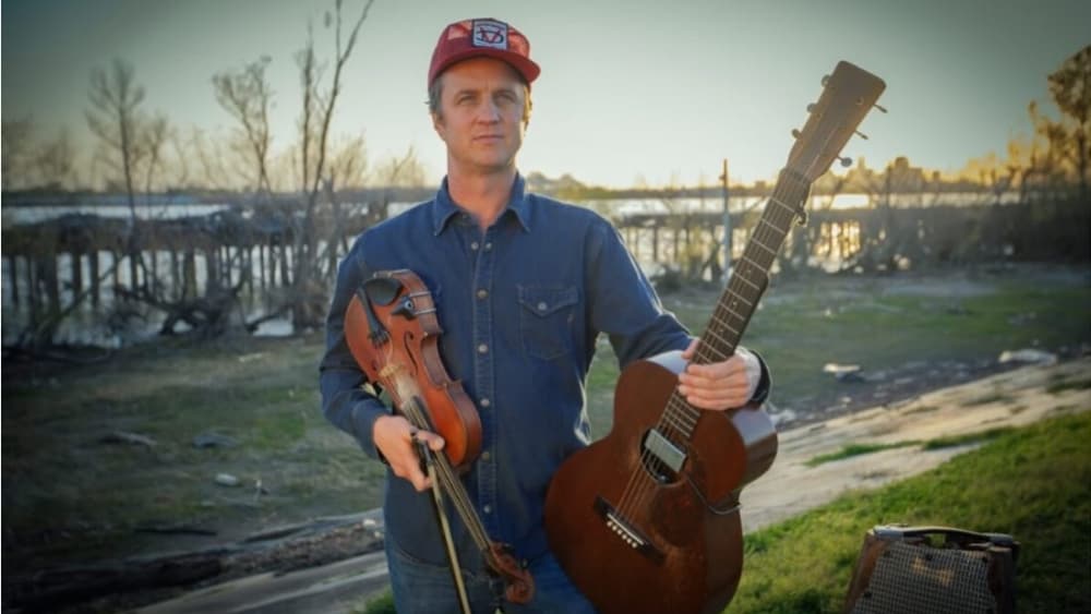 Un homme debout à l'extérieur, un chapeau rouge sur la tête, tenant un violon dans une main et une guitare acoustique dans l'autre.