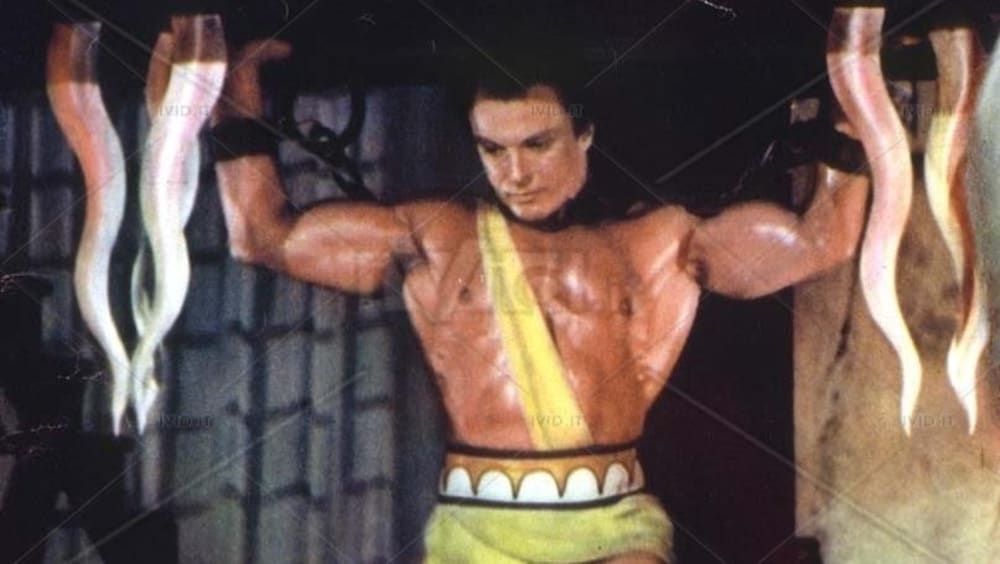 Un homme fort, en pagne jaune, porte une grille à laquelle il est enchaîné au dessus de sa tête.