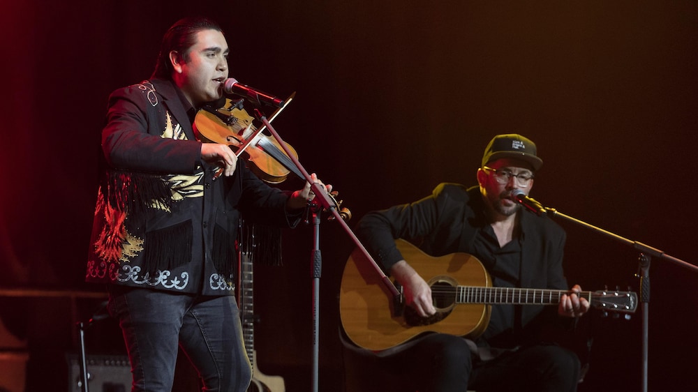 Un homme debout sur scène jouant du violon et un autre assis jouant de la guitare.