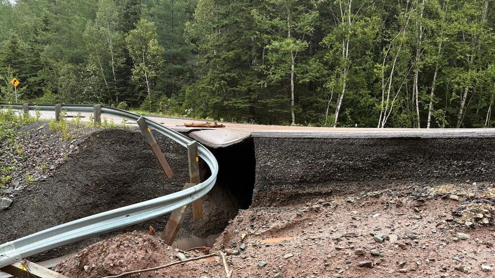 Une route asphaltée traversant une forêt est totalement effondrée et coupée en deux par une profonde crevasse causée par des inondations, laissant voir une glissière de sécurité déformée suspendue au-dessus du vide