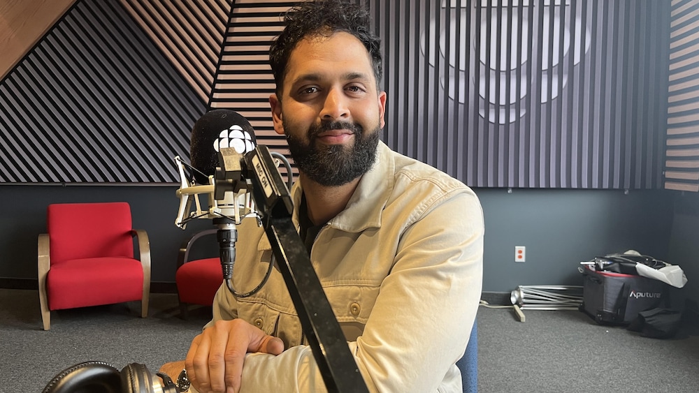 Dans un studio de radio, un homme, vêtu d'une veste beige, sourit devant un microphone.