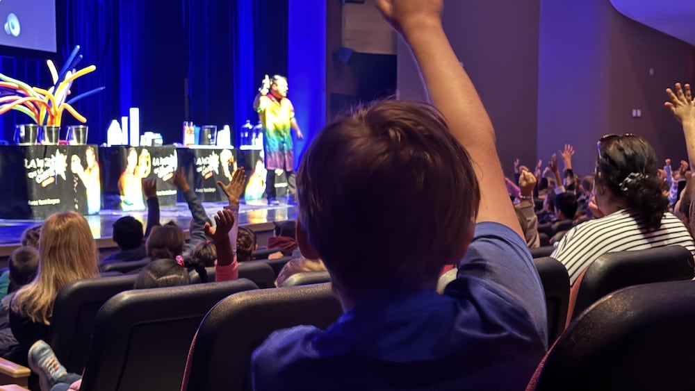 Gros plan sur un jeune enfant levant la main, vu de dos. Il est assis dans une salle de spectacle et entouré d'autres enfants. Sur scène, le chimiste Yannick Bergeron anime un spectacle de science.