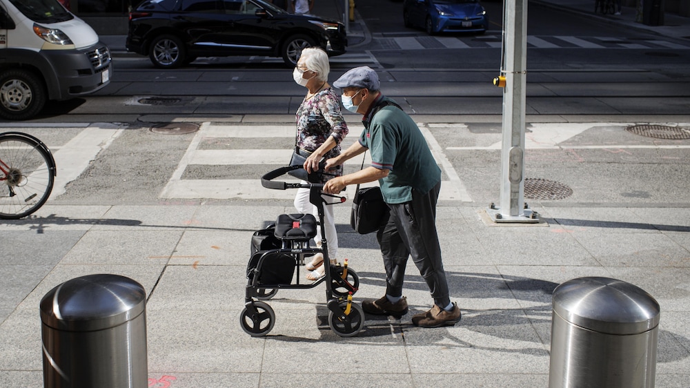 Un couple de personnes âgées portent des masques. L'un d'eux utilisent une marchette. Ils marchent dans une rue de Toronto.