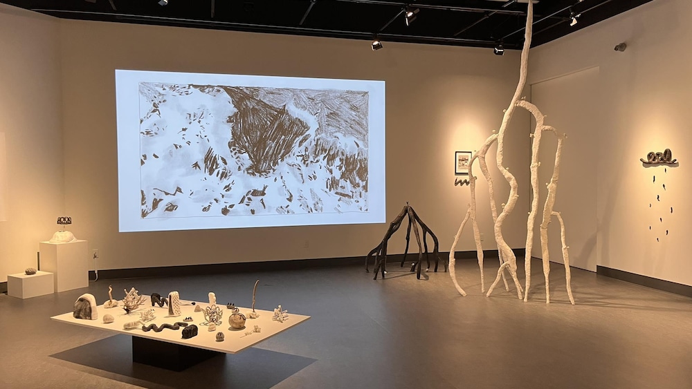 Diverses sculptures blanches et noires occupent une salle de galerie devant une projection murale d’un dessin abstrait.
