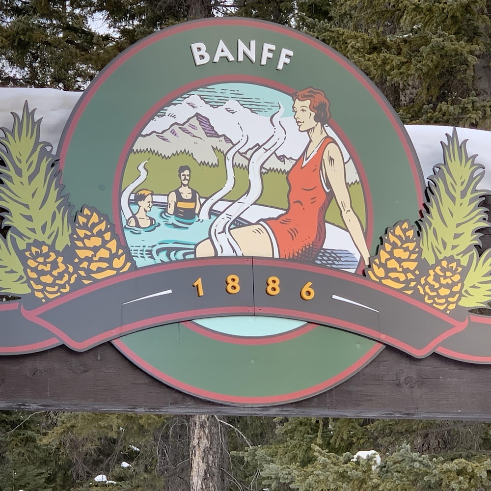 Une affiche en bois indique le nom de Banff et la date 1886 avec une femme en maillot de bain d'époque les pieds dans l'eau. 