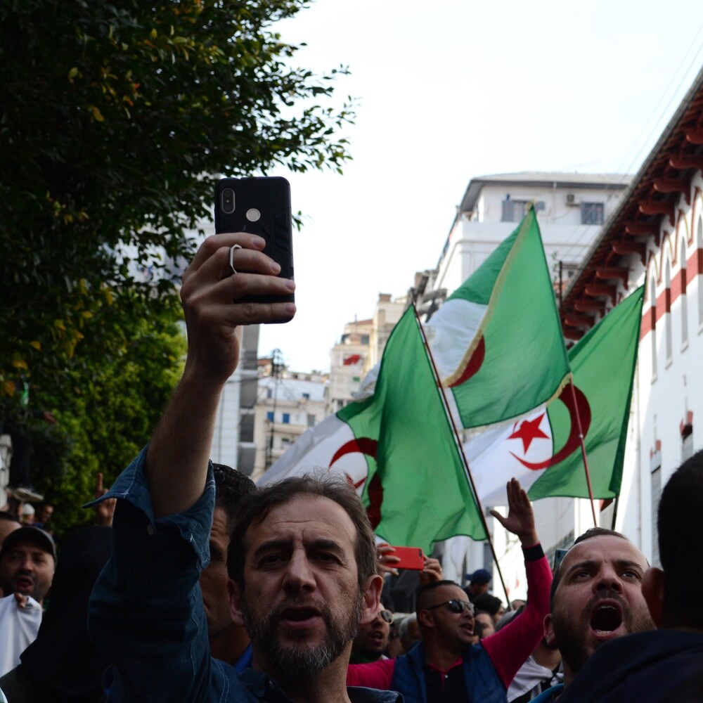 Des manifestants à Alger le 13 mars 2020. Un homme tient un téléphone cellulaire et d’autres brandissent des drapeaux algériens.