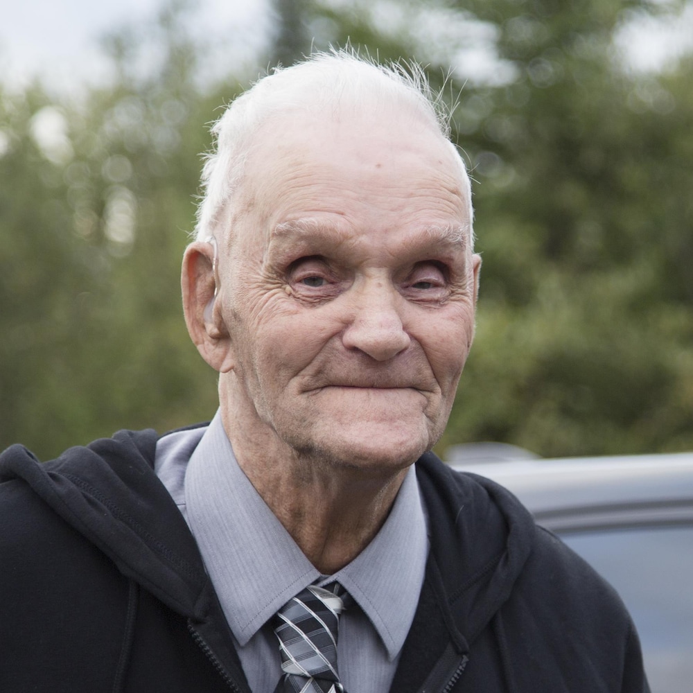 Un homme âgé aux cheveux blancs, debout à l'extérieur.