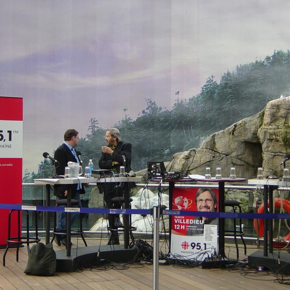 Table avec console d'enregistrement, installée devant un décor du Biodôme de Montréal, deux hommes discutent debout.
