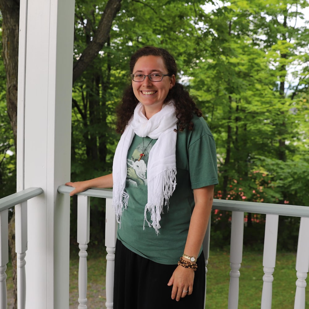 Sœur Sara est vêtue d'un chandail à manches courtes vert avec une illustration de brebis et un foulard blanc. Elle pose pour la caméra sur la galerie d'une maison.