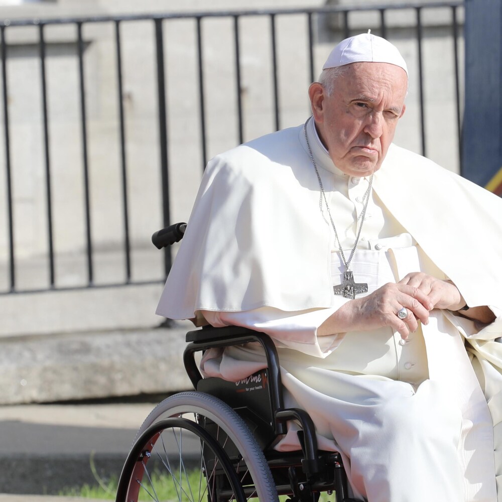 Le pape est vêtu d'une toge blanche et d'un crucifix. Il est assis dans une chaise roulante.