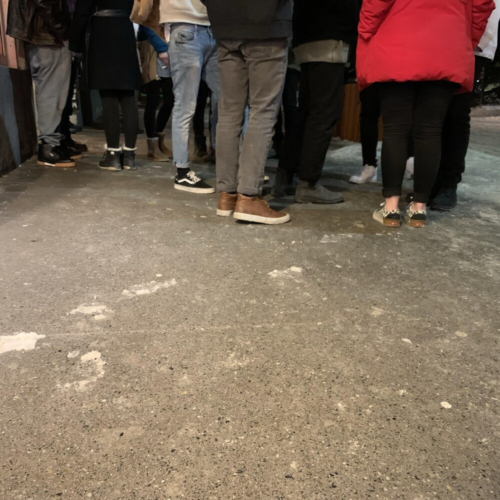 La photo montre une dizaine de jambes avec des chaussures et des bottes aux pieds de gens qui sont debout sur un trottoir en hiver. 