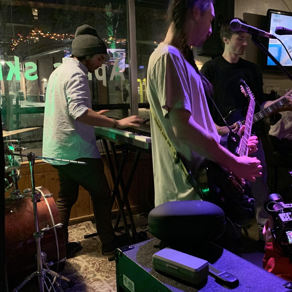 Trois jeunes hommes dont deux avec des guitares et un au clavier font un concert de musique dans un bar sombre. 