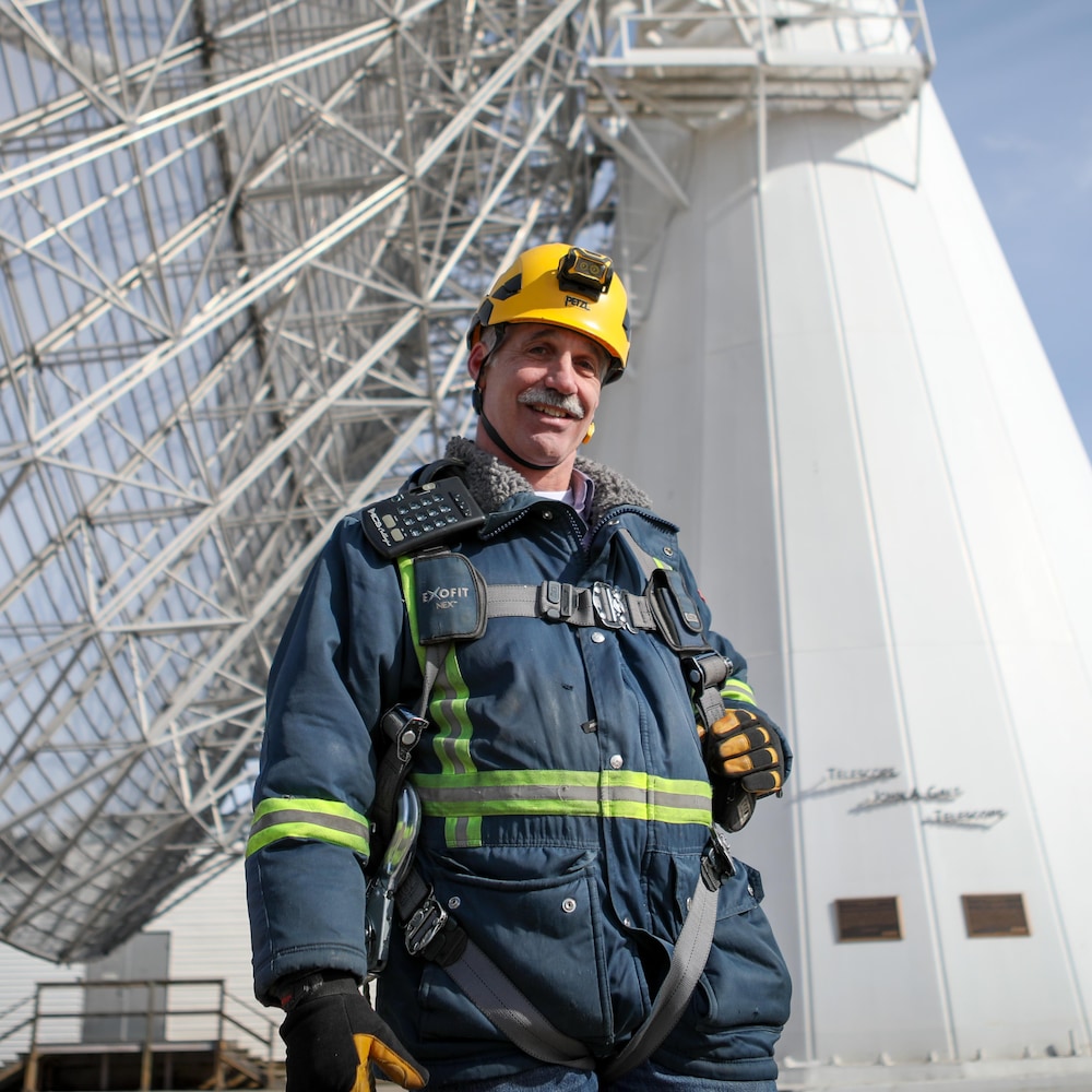 Benoît Robert en bleu de travail, casque sur la tête, se tient devant un radiotélescope de 26 mètres, à l’Observatoire fédéral de Radioastrophysique près de Penticton, en février 2024.