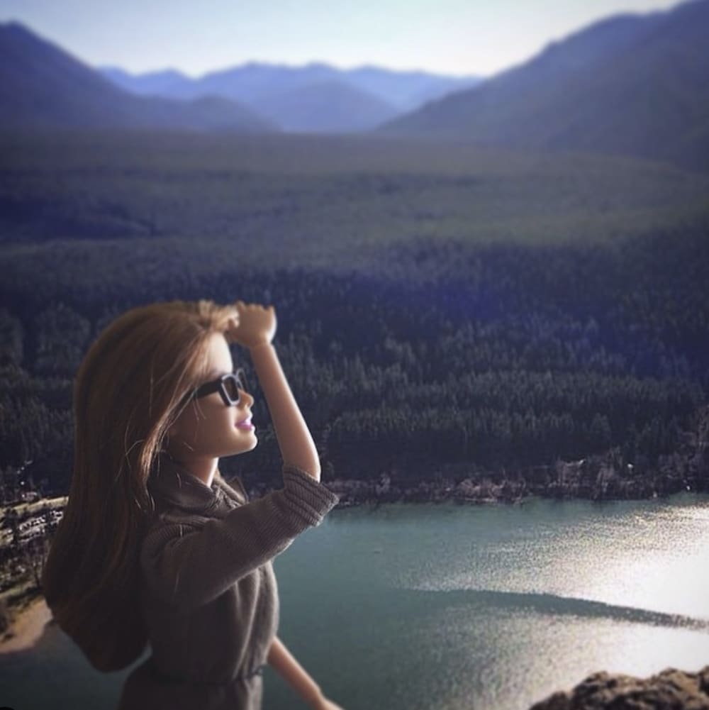 Un montage photo d'une barbie qui regarde au loin du haut d'une montagne avec vue sur un lac.