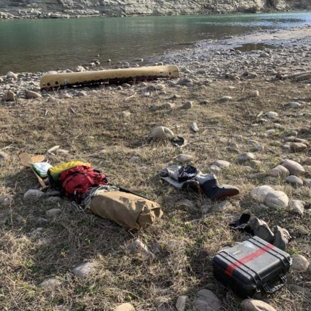 Des sacs étanches, un boîtier et  un canot son éparpillés sur la rive d'une rivière.