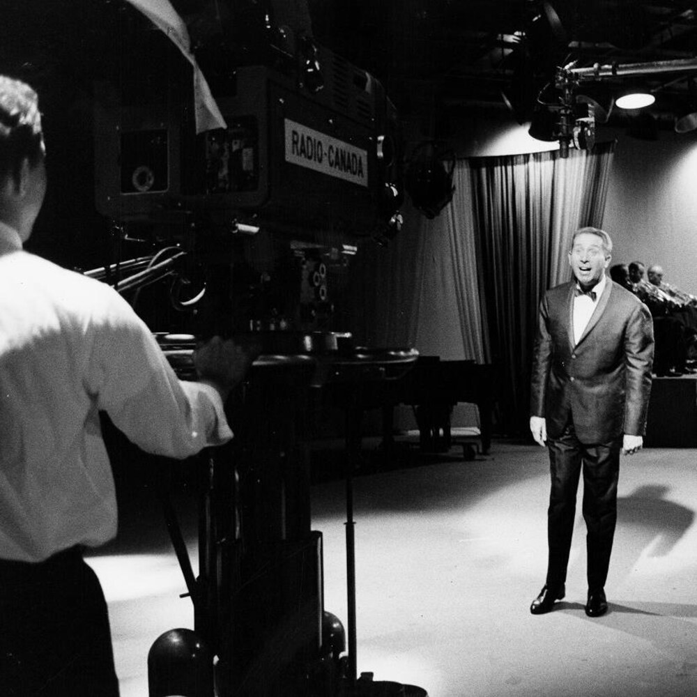 Sur le plateau de l'émission, un caméraman film le chanteur Charles Trenet et derrière, l'orchestre.
