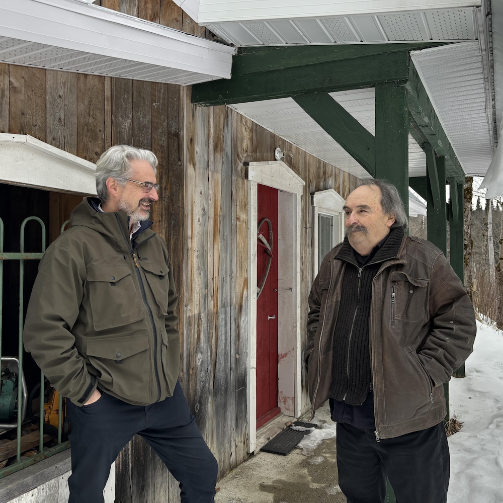 Claude Lepage et Réjean Bilodeau discutent devant une cabane en bois.