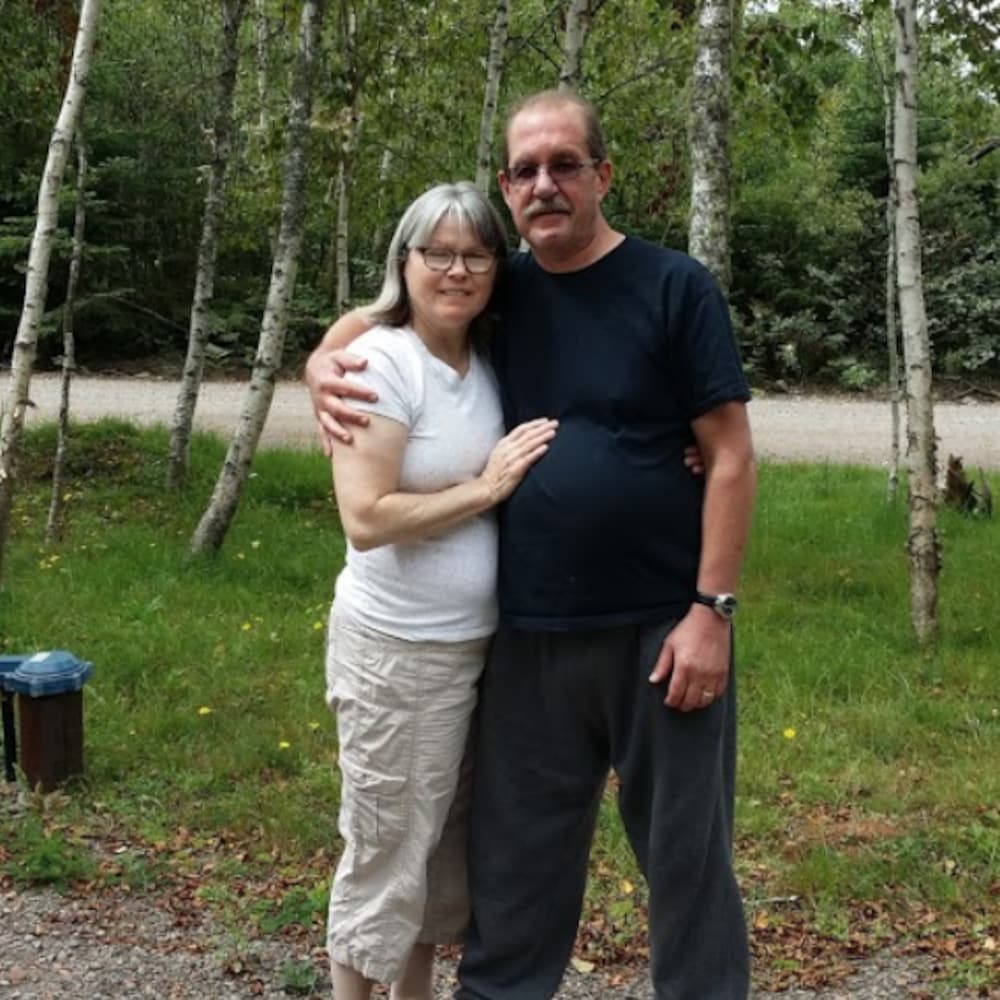 Frank entoure Dawn de son bras pour prendre une pose devant la caméra. Ils se trouvent devant une forêt pour la photo. 