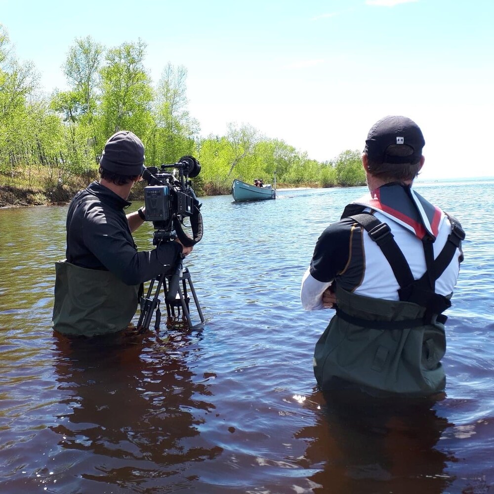 Caméramen avec sa caméra sur pied filme une chaloupe au loin dans le lac un journaliste vu de dos regarde la scène.