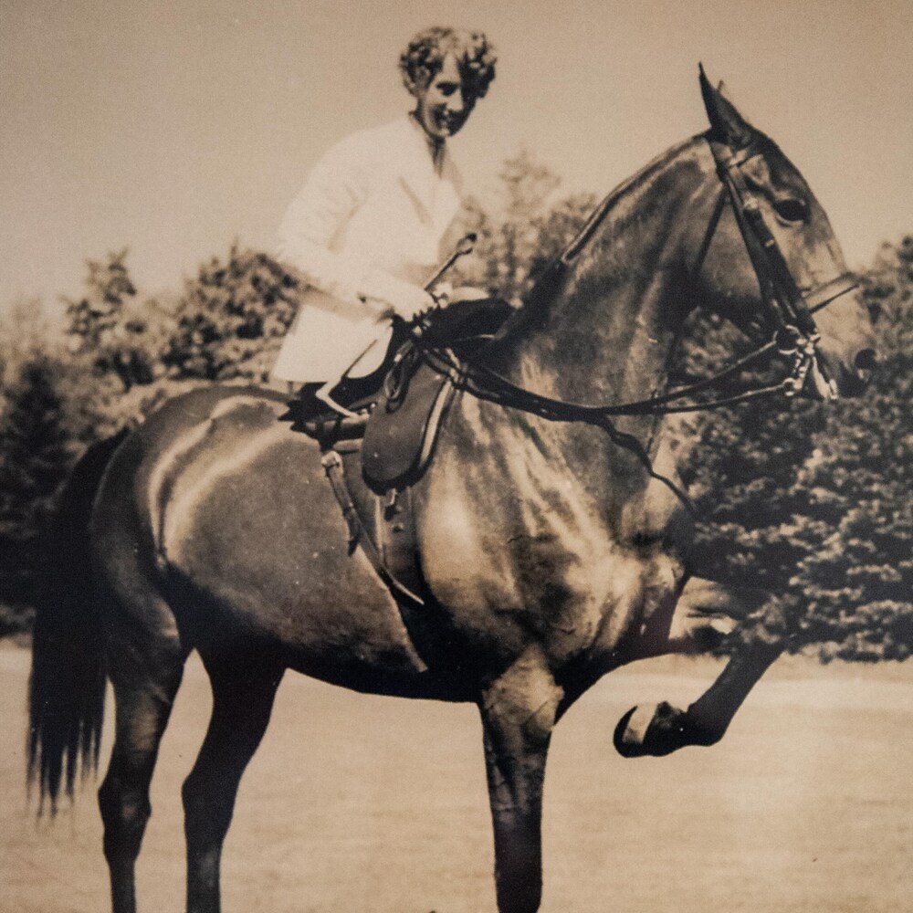 Agnes Slayden sourie pour la caméra tandis que son cheval prend la pose, une patte levée dans les airs. Elle est assise en marquise sur sa scelle.