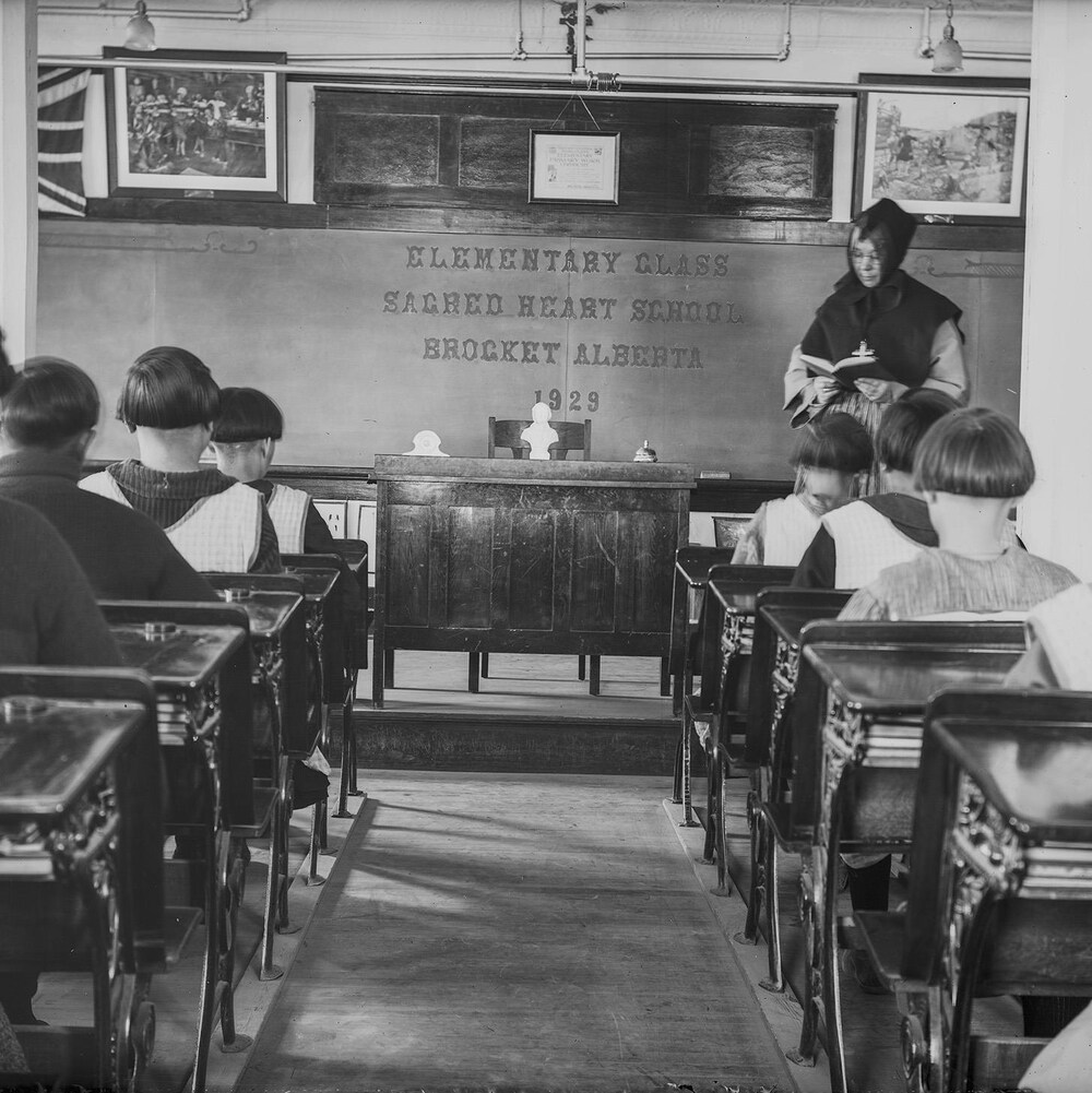 Des élèves, garçons et filles, sont assis à des pupitres (de dos) devant une religieuse qui tient un livre ouvert à l'avant de la classe.