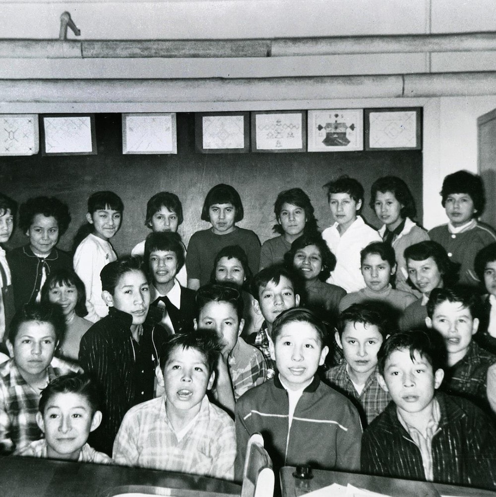 Des élèves, garçons et filles, posent pour la photo dans une classe. Ils sont debout, derrière une rangée de pupitres, et des dessins (motifs autochtones?) sont accrochés au mur derrière le groupe.