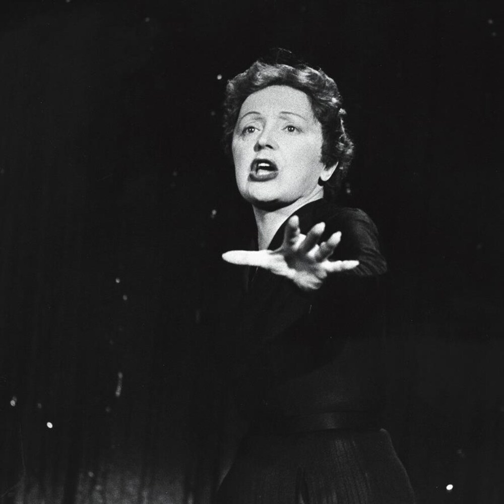 La chanteuse Édith Piaf, en performance, une main tendue devant elle.