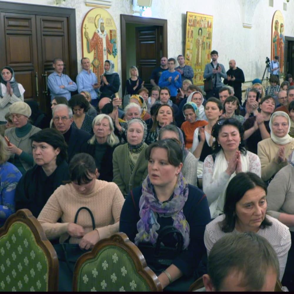 Des hommes et des femmes sont assis en assemblée dans le hall d'une église. Des imageries orthodoxes sont accrochées au mur au fond de la pièce.