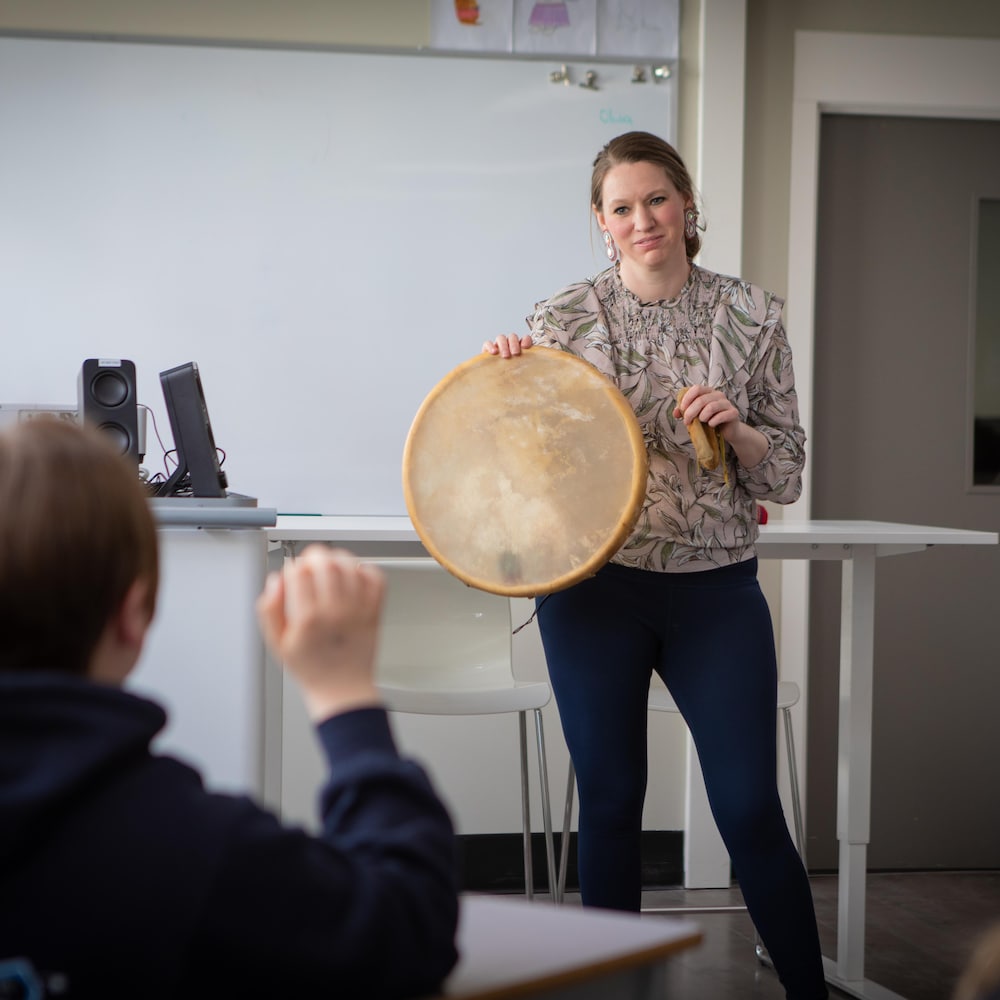 Bonnie Lépine est debout devant une classe et leur montre un tambour traditionnel autochtone. 