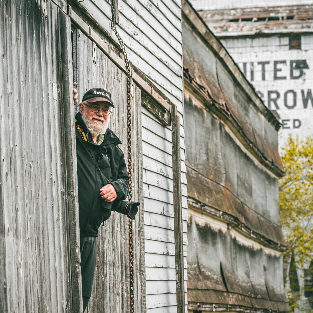 Gordon Goldsborough passe la tête par la porte d'un très vieux silo à grains, son appareil photo en bandoulière, à Barnsley au Manitoba, début juin 2022.