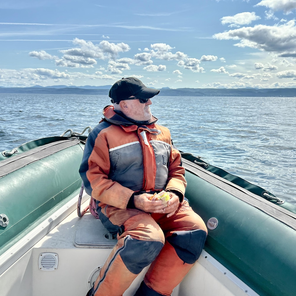 Un homme âgé assis dans un bateau pneumatique qui scrute l'horizon avec un sandwich à la main.